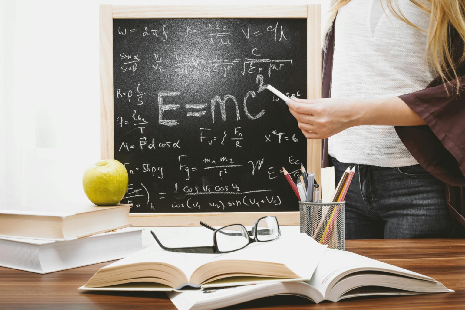 Woman writing physics equations on a blackboard with books and an apple on the desk.