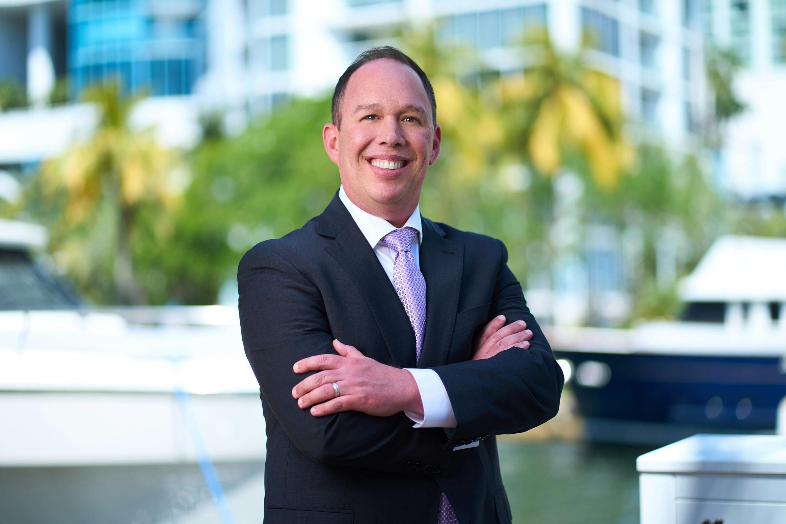 Smiling businessman in a suit standing by a marina with yachts in the background.