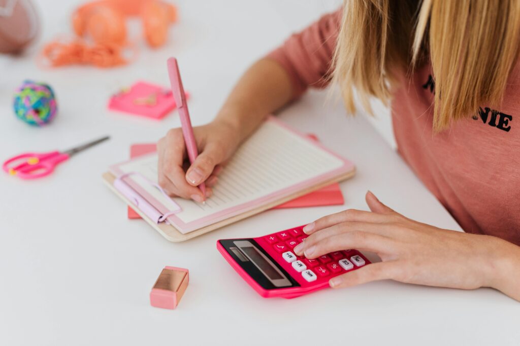 A young student uses a pink calculator while writing notes in a notebook, surrounded by colorful stationery.