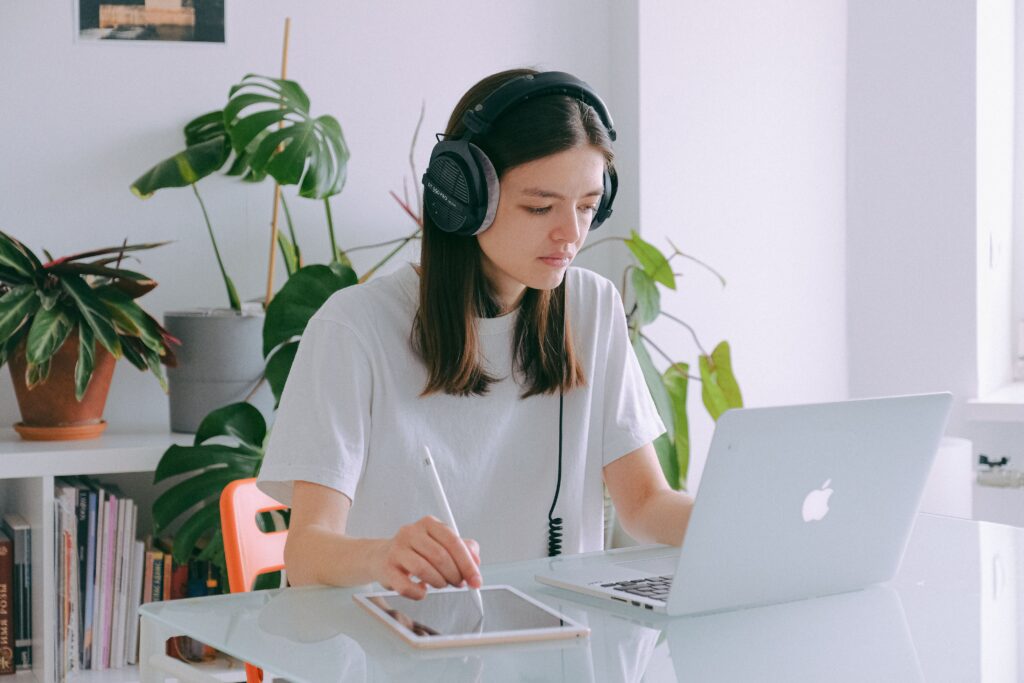 Young woman in home office using a laptop and drawing tablet, wearing headphones, illustrating remote work.